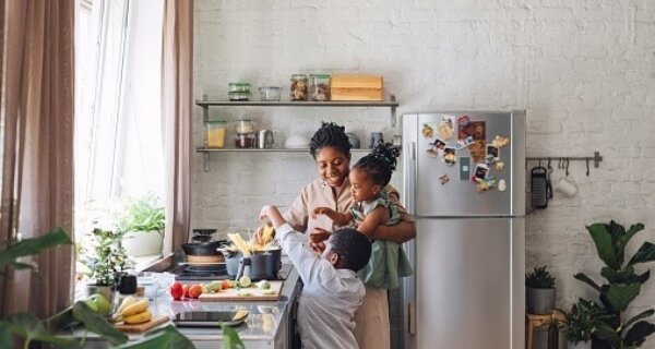 Bright kitchen with a parent and two children at a counter, preparing food, magnets on the fridge, open shelves, and potted plants by a sunlit window.