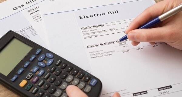 Close-up of a hand holding a blue pen over an electric bill; a calculator and a gas bill sit nearby on a desk.