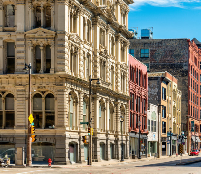 Sunlit city street with a grand beige stone building on the left and colorful brick storefronts along the right, with street lamps and parked vehicles.