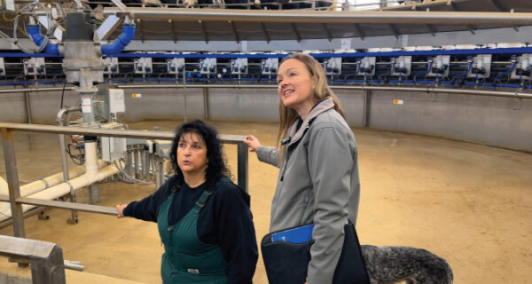 Two people stand by a railing in an industrial facility, one wearing green overalls and a dark shirt, the other in a gray jacket with a blue folder, looking upward.