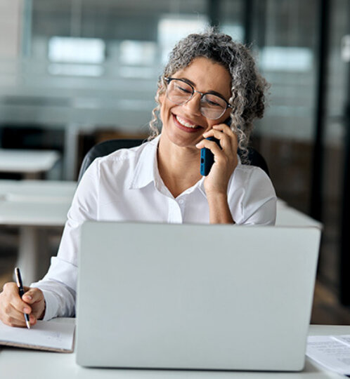 Person with gray curly hair and glasses sits at a desk with a laptop, talking on a cellphone and taking notes in a bright office.