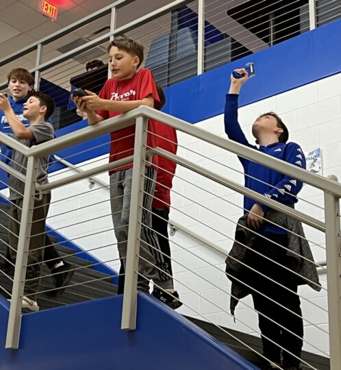 Four children stand on a stair landing in a school, filming with smartphones; one raises a phone toward the ceiling while leaning on the metal railing.