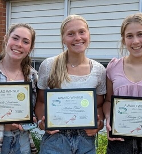 Three people stand outside a brick building, each holding a framed award and smiling; left in plaid, middle in white, right in pink.
