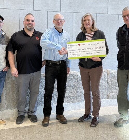 Five people pose indoors against a stone wall; one holds a large energy-efficiency grant check to the School District of Arcadia while a handshake occurs.