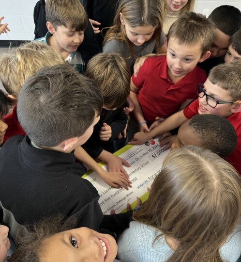 A diverse group of schoolchildren crowds around a large ceremonial check for $2,500, with a smiling child in the foreground looking up at the camera.