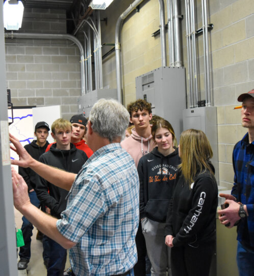 An older person in a plaid shirt demonstrates a control panel to a group of students in an industrial room with exposed wiring and concrete walls.