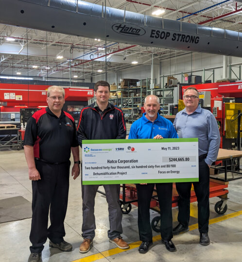 Four people stand in an industrial workshop, holding a large ceremonial check from Hatco Corporation for a dehumidification project, with machinery and red tool cabinets in the background.