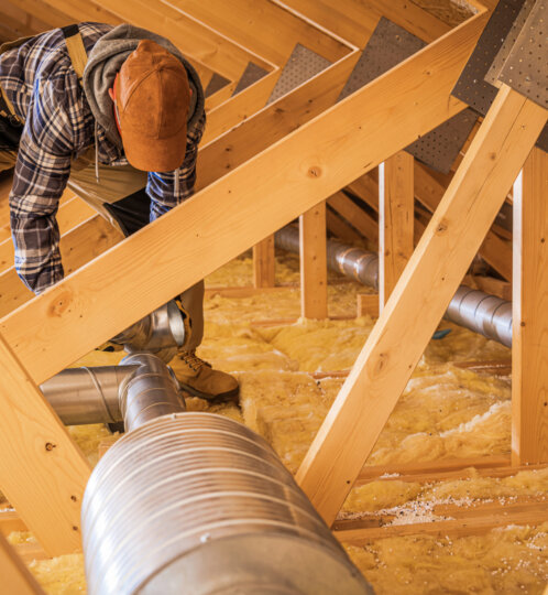 A person in a plaid shirt and orange cap kneels in an unfinished attic, adjusting silver ductwork among wooden framing and yellow insulation.
