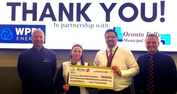 Four people stand in front of a screen that says THANK YOU!, with two holding a large ceremonial check and everyone smiling.