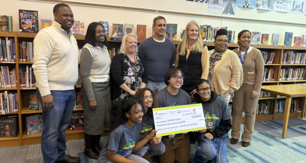Diverse group of adults and kids pose in a library, some standing and some kneeling, holding a large ceremonial check in front of bookshelves.