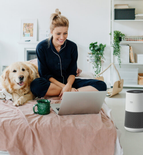 Person in dark pajamas sits cross-legged on a pink bed, smiling at a laptop; a golden retriever lies beside with a green mug and a white speaker nearby.