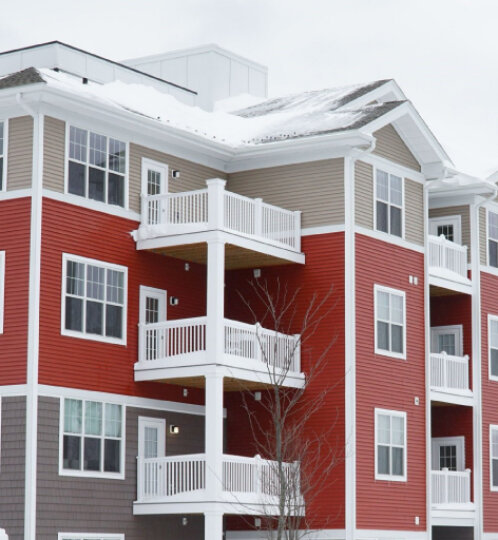 Multistory red and tan apartment building with white balconies, snow on the roof, and a bare tree in front.