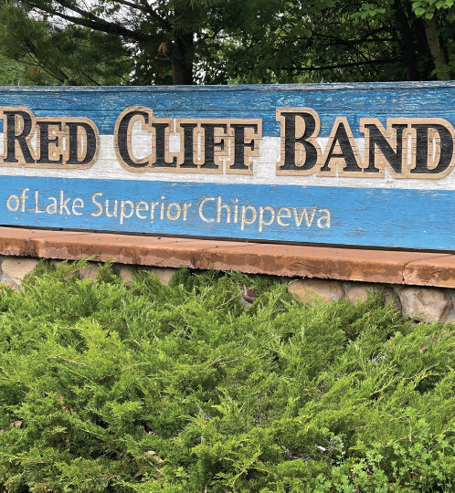 Blue weathered wooden sign reading Red Cliff Band of Lake Superior Chippewa, mounted on a stone base with green shrubs in front and trees behind.