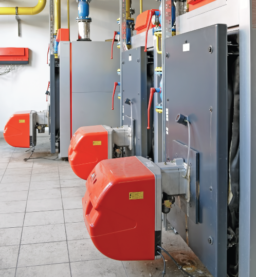 Industrial boiler room with gray control cabinets and red motorized valve units, blue and yellow piping along a tiled floor.