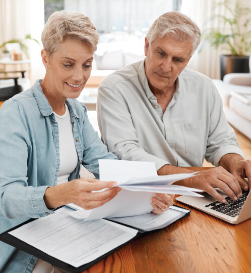 Two adults review documents at a wooden table with papers, a binder, and a laptop in a bright living room.