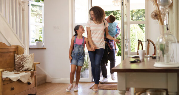 Bright home entry with three people entering through open doors: an adult with a striped shirt and bag, a girl in denim overalls, and another person carrying a child on their back.
