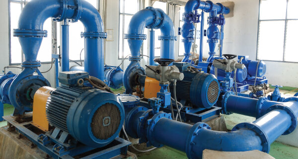 Industrial mechanical room with multiple blue pumps, motors, and interconnected piping; metal components mounted on a concrete floor near large windows.