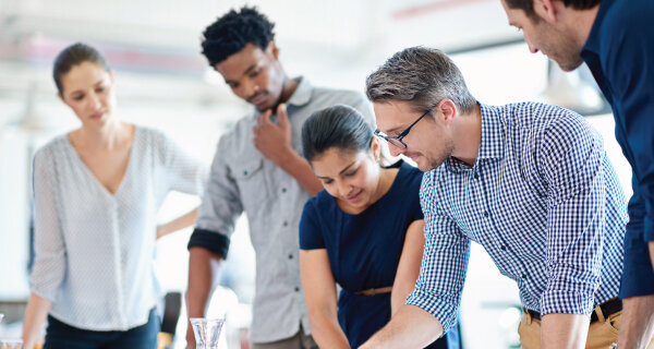 Five diverse professionals collaborate around a table in a bright office, examining a document as they discuss, with a glass of water nearby.