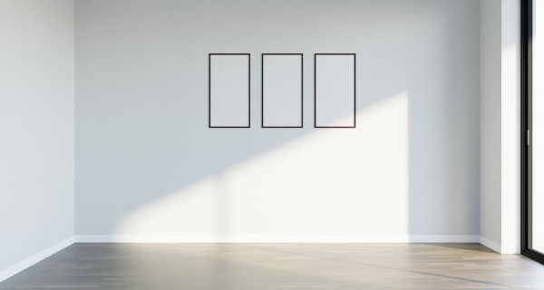 Bright minimalist room with white walls and a wooden floor; three tall empty frames hang in a row, while sunlight casts a diagonal shadow from a large window on the right.
