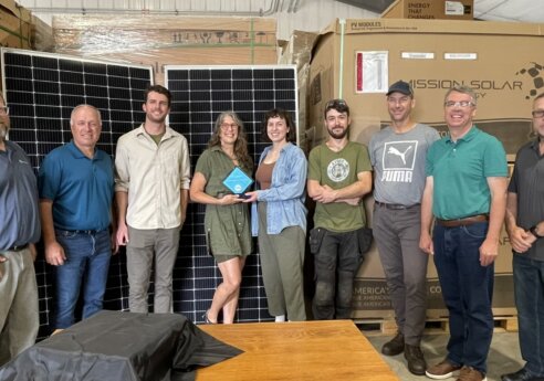 Group of people in a warehouse with large solar panels and boxed solar equipment; two center people hold a blue diamond award.