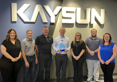 Seven people stand in an office lobby under a large KAYSUN sign; the center person holds a blue award while others smile.
