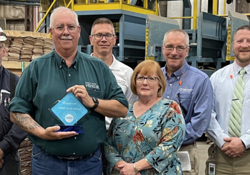 Group of professionals posing in a veneer warehouse; center person holds a blue award plaque, with stacks of plywood and shelving in the background.