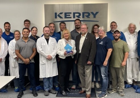 Group of staff posing in a Kerry facility in front of a large Kerry sign; several wear white lab coats and blue shirts, one person holds a blue award plaque.