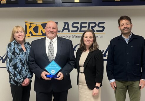 Four people stand indoors in front of a KM Lasers sign; the central person in a dark suit holds a blue diamond-shaped trophy marked 2023, flanked by three others.