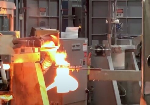 Person wearing a bandana and gloves stands beside a molten metal forge, with bright orange flames and industrial machinery in a factory setting.