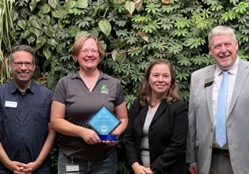 Seven people in business casual attire stand in an office hallway in front of a leafy green wall; one person at center holds a blue-tinted award.