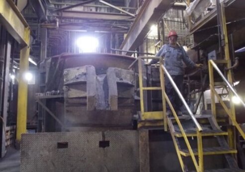 A person in a red hard hat stands on a yellow-railed stair platform beside a large dusty industrial machine in a factory.