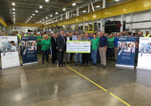 Group of people in a factory posing with a large ceremonial check and Alliance banners, yellow crane overhead and equipment in background.