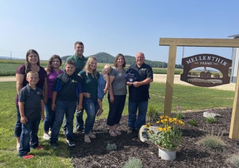 Group of people posing outdoors beside a wooden Mellenthin Farms sign; one person holds a plaque, with flowers and fields in the background.
