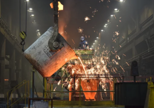 Foundry interior with a crane bucket pouring molten metal, sparks flying as a worker in protective gear stands on a platform above.