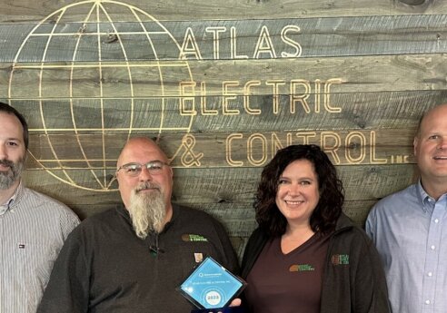 Six professionals stand in front of a rustic wood wall with Atlas Electric & Control branding, smiling; one holds a blue award plaque.