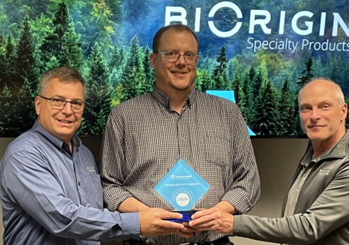 Three people indoors, smiling, jointly holding a blue diamond-shaped award marked 2024, with a forest-themed screen and company branding in the background.