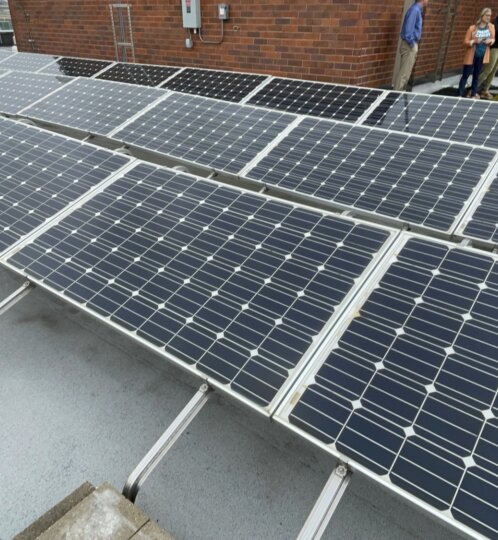 Rooftop solar array with rows of dark panels on metal rails and ballast blocks; several people stand near a brick building in the background.