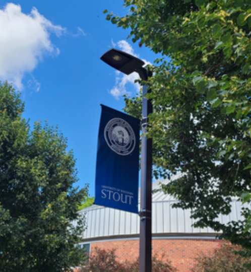Blue banner with a white circular seal and the word STOUT hanging from a black street lamp, flanked by green trees and a brick building under a bright blue sky.