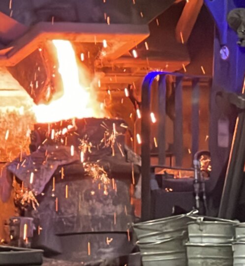 Industrial foundry scene with molten metal pouring from a ladle into a mold, sparks flying, heavy machinery, and a blue-lit workshop.