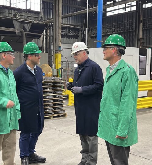 Group of workers in a factory wearing green and white hard hats, safety glasses and gloves, standing in a semicircle near machines and yellow barriers