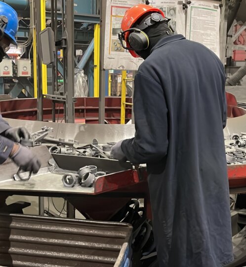 Two workers in hard hats and protective gear sort metal clamps on a circular tray in a busy factory.