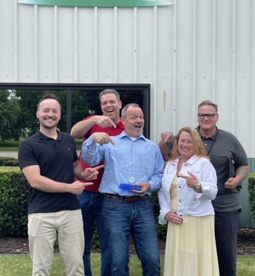 Five people stand outside a light gray metal building, smiling. A person in a blue shirt holds a blue award while others point and cheer.