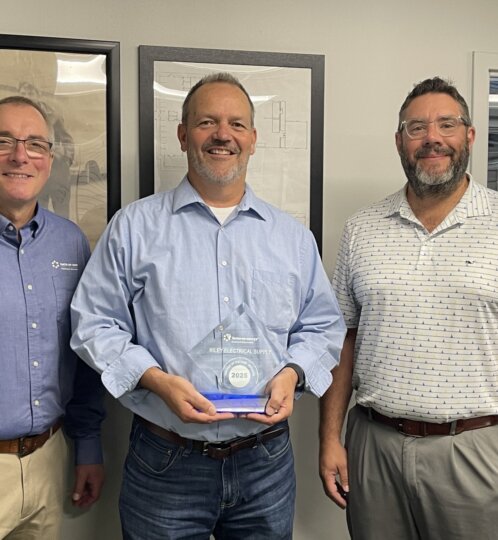 Three people stand in an office, smiling; the center person holds a glass award labeled 2025, flanked by two coworkers in casual shirts.