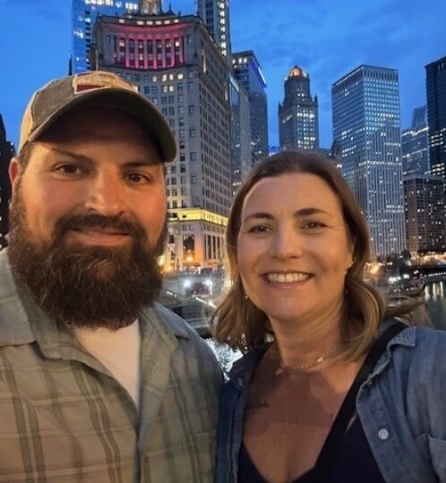 Two people take a selfie by a riverfront cityscape at dusk; one with a beard and cap, the other in a denim shirt, both smiling with illuminated skyscrapers behind.