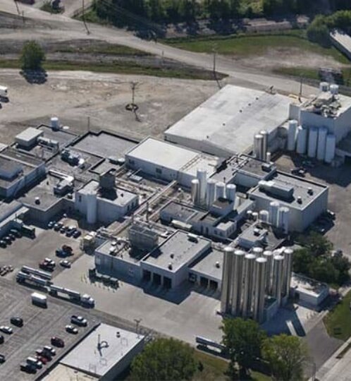 Aerial view of a large industrial facility with white silos, flat-roofed buildings, loading areas, and a spacious parking lot.