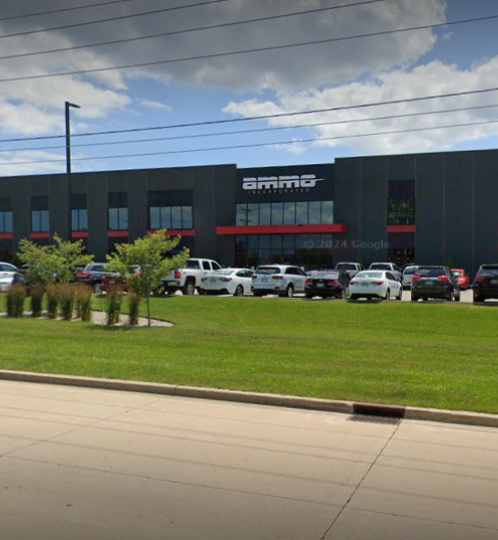 A modern dark-gray office building with red trim sits behind a long parking lot full of cars, with green lawns, small trees, and flagpoles under a blue, partly cloudy sky.