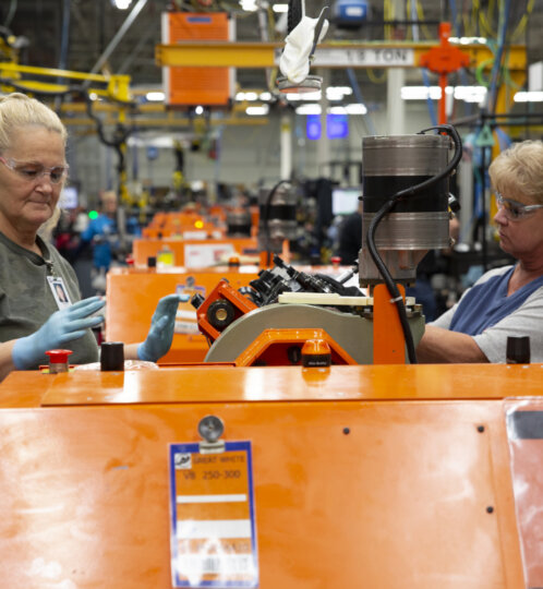 Two workers wearing safety glasses and gloves operate an orange industrial machine on a busy factory floor.