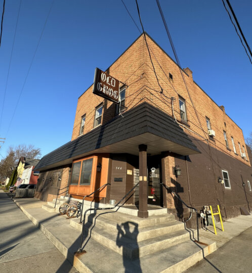 Two-story brick corner building with a dark awning and "OLD GROG" sign; stairs to the door, a bicycle nearby, and power lines overhead.