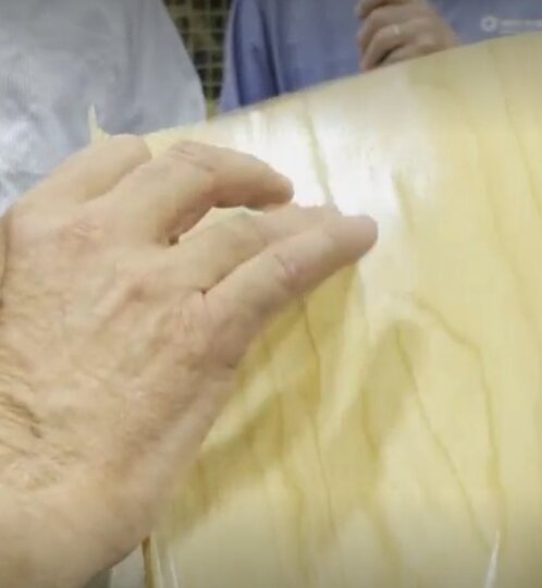 Close-up of a hand smoothing pale wood veneer onto a curved surface, with two people visible in the background.