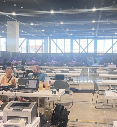 Spacious hall with rows of tables and laptops; three people work at a foreground table with drinks, while signs E8–E11 and a black curtained backdrop are visible.